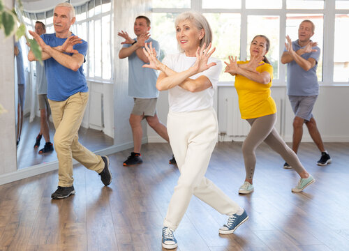 Expressive cheerful woman participating in upbeat Zumba class for group of seniors in mirrored fitness studio..