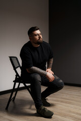 A stylish man with tattoos and glasses sits contemplatively in a chair, exuding a calm and...