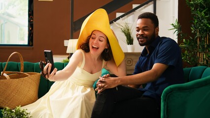 Tourists using videocall conference in lounge area, talking to family after they arrive on summer vacation. Diverse couple enjoying teleconference chat online, luxury hotel lobby. Handheld shot.