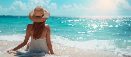A woman in a white sundress and straw hat sits on a sunlit beach, facing the sparkling turquoise sea. Ample room for copyspace - obrazy, fototapety, plakaty