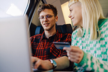 Young couple sitting in train and using credit card and laptop.