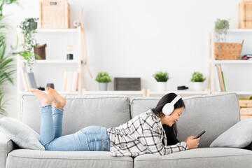 Chinese woman lying on the sofa using phone and listening to music with headphones