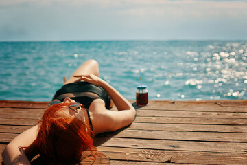 Red-haired woman is enjoying a vacation at the sea. Female in a swimsuit is lying on a wooden pier with a bottle of lemonade and sunbathing. Summer trip. Copy space for travel agency advertising.