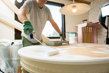 Middle-age man painting old wooden table with paint roller in white color. Furniture restoration,...