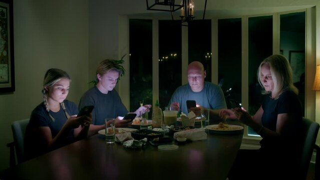 A family of four look at their smartphones in a dark room while a man binge eats his dinner