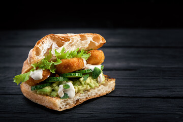 Appetizing sandwich on a wooden board. Baguette sandwich with filling from lettuce, slices tomato. Dark wooden background. View from above. Close-up. Macro photography.