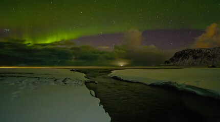 Snowy winter landscape where stream meets the sea under the cloudy night sky with northern lights.