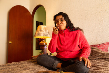 A woman sits pensively on a neatly made bed, her fingers delicately holding a joint to her lips