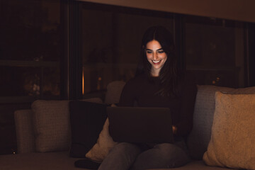 Close up of one young happy cheerful woman smiling and using laptop computer at home sitting on sofa working and studying alone at late night. Millennial girl surfing the net indoor with light screen