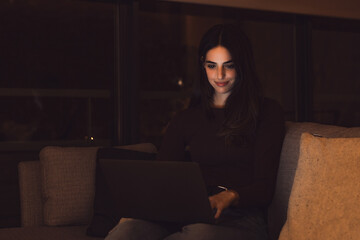 Close up of one young happy cheerful woman smiling and using laptop computer at home sitting on sofa working and studying alone at late night. Millennial girl surfing the net indoor with light screen