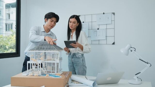 Beautiful young caucasian project manager brief asian engineer by using tablet while engineer measuring house model on meeting table with blueprint, architectural equipment scatter around. Immaculate