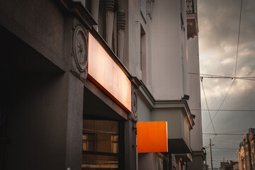 Empty orange square shop signboard hanging on a building facade, visible against a cloudy sky in a...