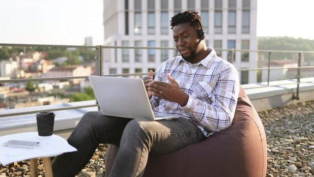Relaxed African American man conducting web conference via laptop and headset while staying on flat roof. Professional entrepreneur working outside of corporate offices via wireless devices.