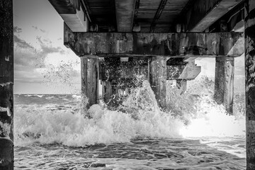 Dynamic monochrome photograph showcasing the power of the ocean as waves crash under a pier, ideal...