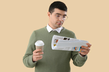 Young man in eyeglasses with cup of coffee reading newspaper on beige background