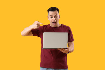 Shocked young man pointing at laptop on yellow background. Deadline concept