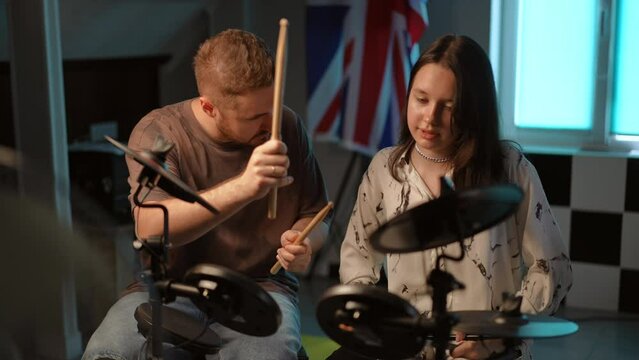 Slow motion. A male teacher explains to his pupil how hard she needs to hit an electronic drum kit with drumsticks while sitting in the studio of a music school