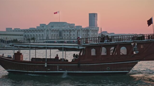 Doha corniche in Doha, Qatar sunset shot showing Dhow in Arabic gulf with Qatar flag waving, cars in the street, people walking and Amiri Diwan in background