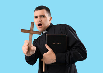 Shocked young priest with Holy Bible and crosses on blue background