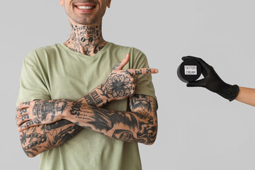 Tattooed man pointing at master holding jar of tattoo cream on grey background