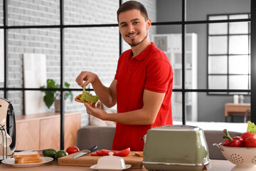 Young man making delicious sandwich with crispy toasts in kitchen - obrazy, fototapety, plakaty