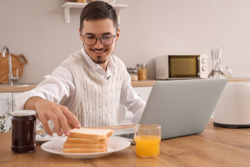 Fototapeta na wymiar Young man with laptop having tasty toasts for breakfast in kitchen
