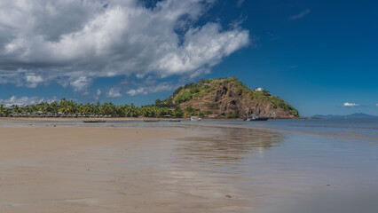 The waves of the ocean spread over the sandy beach. Boats are visible in the distance, a row of sun loungers under sun umbrellas. A hill against a blue sky and clouds. Reflection on wet sand. Nosy Be