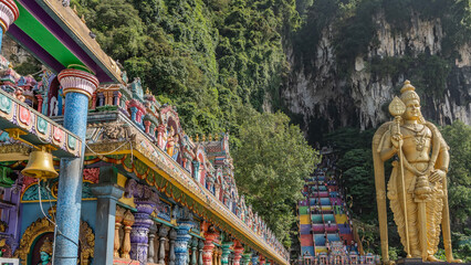 The famous Batu caves. The Hindu temple is decorated with sculptures of gods. People climb the rainbow colored stairs. A huge golden statue of the god Murugan on a rock background. Kuala Lumpur.