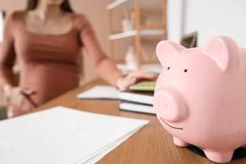 Piggy bank on table of pregnant woman at home, closeup. Maternity Benefit concept
