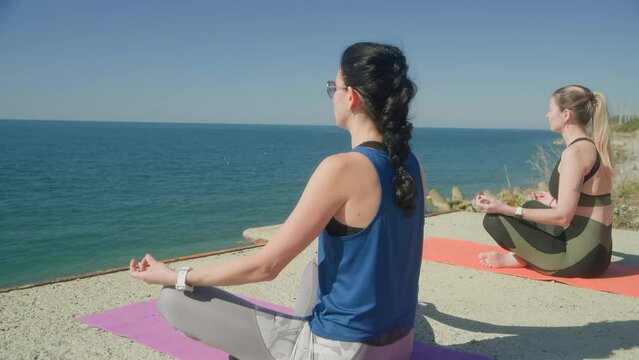 Women meditating sitting in lotus pose on yoga mats by sea admiring seascape. Females practicing yoga on wild nature in sunny day. Sport activity recreation mental spirit practice inner life concept.