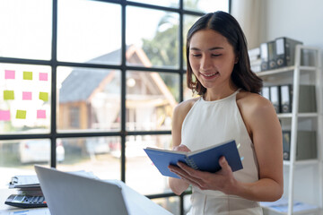 Beautiful Asian businesswoman working on a laptop and having fun at work and reading review book documenting detailed financial information earning peace of mind.