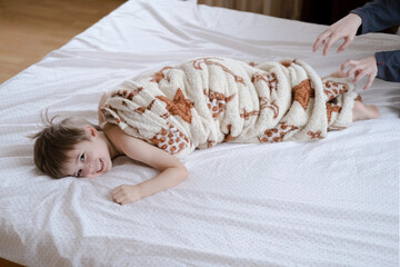 Child toddler boy is wrapped in a blanket like a roll, his head sticks out of the blanket. He is laughing, having fun on the bed. Hands is tickling. - obrazy, fototapety, plakaty