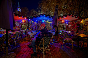 A woman sits in the evening in an empty street cafe illuminated with lights and garlands.