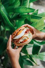 People's hands are holding burger bagels filled with vegetables, chicken, eggs, behind there are plants