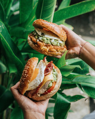 People's hands are holding two burger bagels filled with eggs, vegetables, chicken, sauce, and cheese, behind are plants