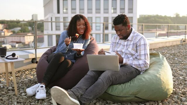 Portrait of happy African colleagues with laptop and bowls of fresh healthy salad spending time together at cozy beanbag during break at rooftop outdoors of office. Business lunch.