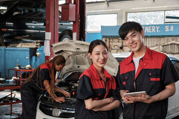 Two Asian professional mechanics, male engineer and partner smile and look at camera, inspect repair work checklist at garage, service car maintenance, and fixing specialist occupations auto industry.