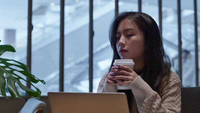 Nostalgic young, pretty Asian woman sits in cafe in front of her laptop, holding a up of coffee, thinking about life.