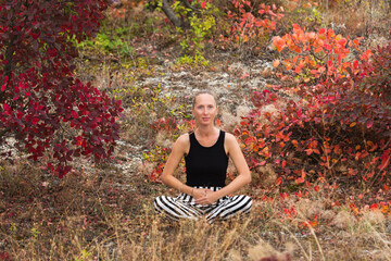 Happy young adult woman sitting in the lotus position against a background of autumn greenery and...