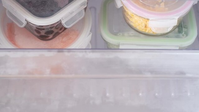 Female hand opens drawer with glass boxes of frozen vegetables in the freezer, top view.