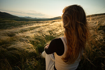 A young adult woman sits in the green grass of golden feather grass and enjoys the warm rays of the...