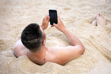 Man lying buried in a sand with smartphone in hands. Holiday and online communication on sea beach
