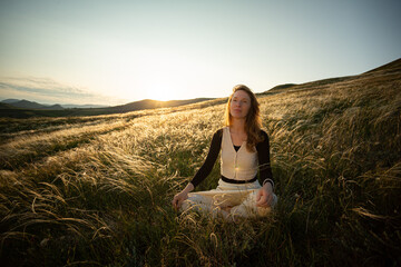 A young adult woman sits in a lotus position in the green grass of golden feather grass and enjoys...