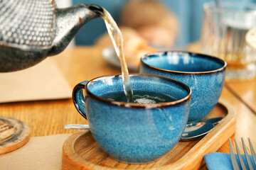 Brewing and Serving Tea. Teapot Pouring Green Tea in Blue Ceramic Cup on Wooden Cafe Table. Two...