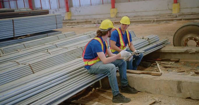 Two builders Construction workers in safety gear sit on steel rods construction materials, engaged in discussion over building plans at a site