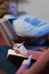 An adult female psychologist interviews a female patient and takes notes in a notebook. The focus is on the doctor's notes, and the patient is in a blur. Vertical composition