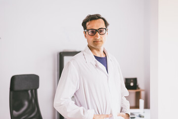 An adult male Caucasian scientist in glasses and a white coat stands and confidently looks forward against the backdrop of the laboratory