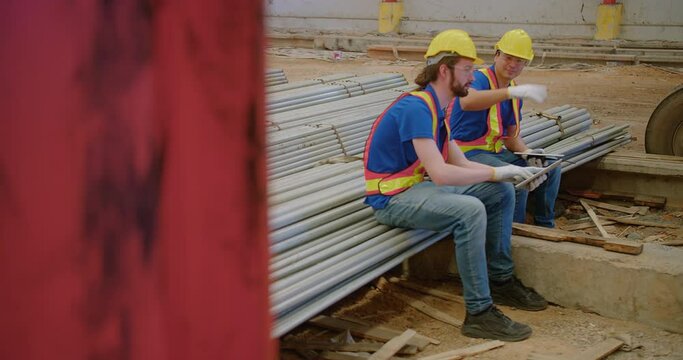 Two builders Construction workers in safety gear sit on steel rods construction materials, engaged in discussion over building plans at a site