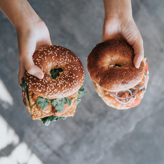 A person's hand holds two burger bagels filled with melted vegetables and cheese