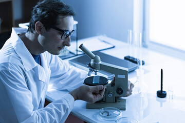 An adult male Caucasian scientist in glasses and a white coat works with a sample under study on a microscope. Color correction - obrazy, fototapety, plakaty
